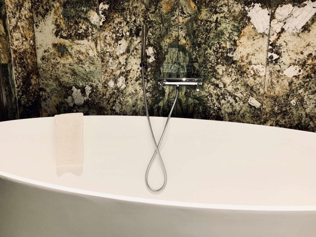 łazienka-w-pokoju-hotel-warszawa-ściany-kamienne-zielony-kamień-do-łazienek-inspiracje-luskusowe-wnętrze-wanna-wolnostojąca-chromowany-prysznic